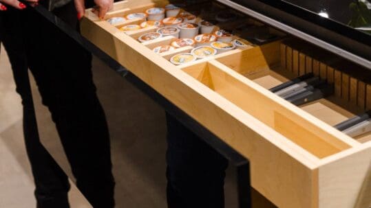 Open wooden pull-out drawer filled with rows of coffee pods under a glass display; person with red nail polish nearby.