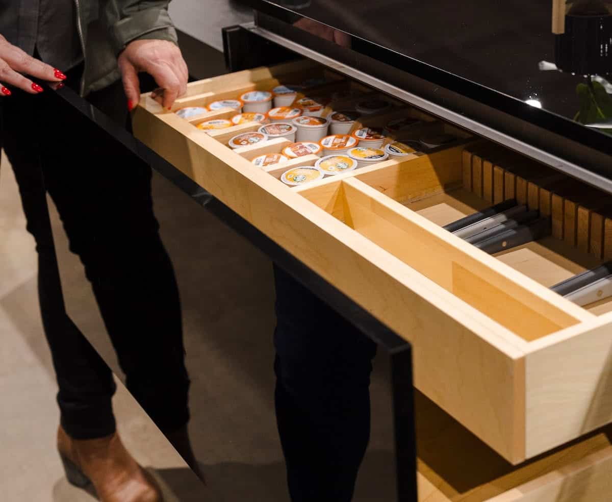 Open wooden pull-out drawer filled with rows of coffee pods under a glass display; person with red nail polish nearby.